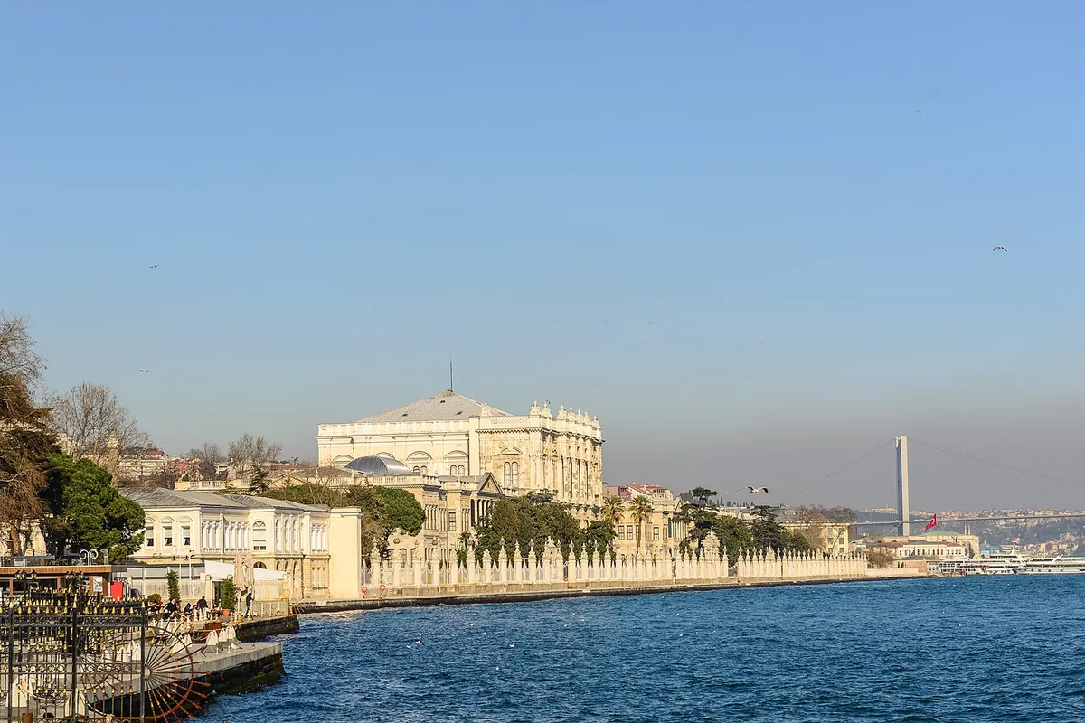 The long cream waterfront of Dolmabahçe Palace beside the Bosphorus, with a mosque in the foreground and the bridge in the distance.