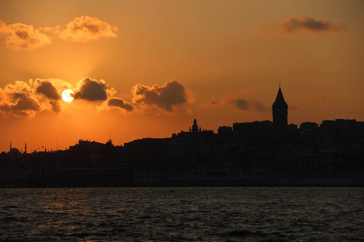 The silhouette of the Galata Tower and the old city against an orange sunset sky above the water.