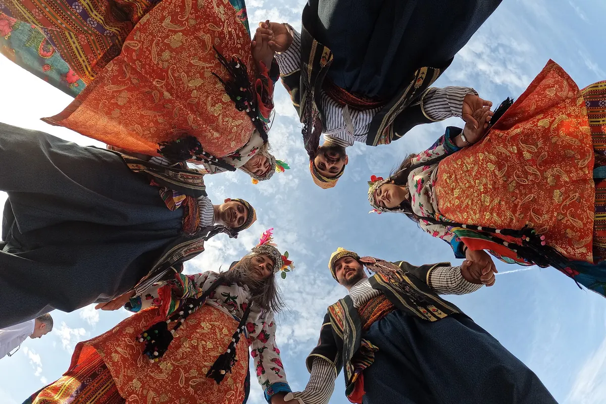 Dancers in embroidered folk costume holding hands in a circle, photographed from below against the sky.