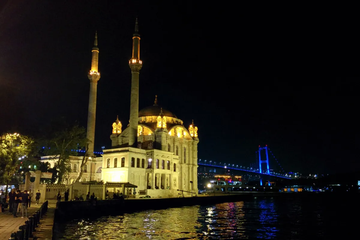 Ortaköy Mosque lit gold at night on the water's edge, with two slim minarets and the blue-lit Bosphorus Bridge behind it.